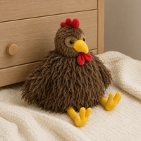 Peluche coq marron à longs poils posée sur une couverture ivoire près d'une commode en bois clair