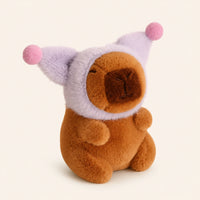 Peluche capybara avec bonnet violet a pompons, environ 12 cm, fond ivoire