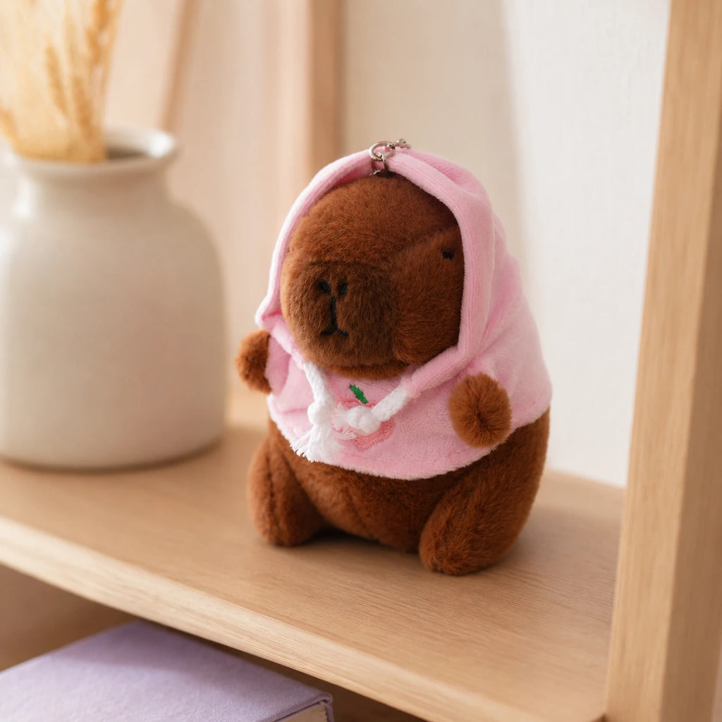 Peluche porte-cles capybara a capuche rose posee sur une etagere, mise en situation