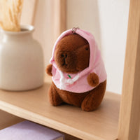 Peluche porte-cles capybara a capuche rose posee sur une etagere, mise en situation