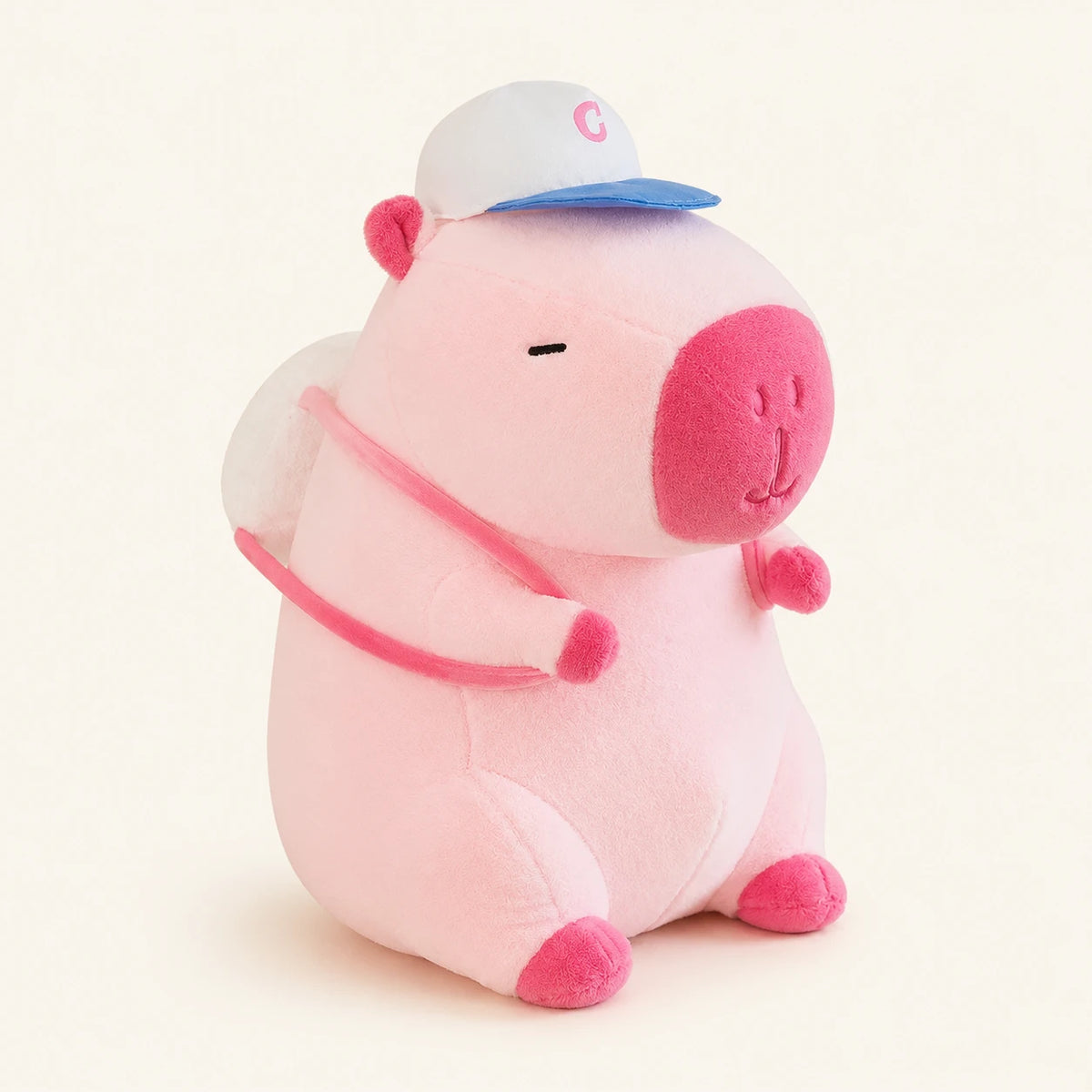 Peluche capybara rose avec casquette et sacoche, vue principale sur fond ivoire