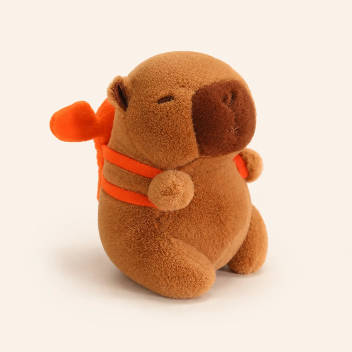 Peluche capybara brun avec petit sac à dos orange, vue de face sur fond ivoire
