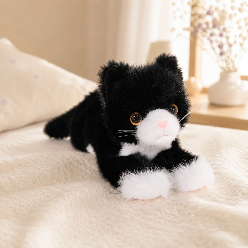 Peluche chat realiste noir et blanc posee sur un lit, mise en situation cosy